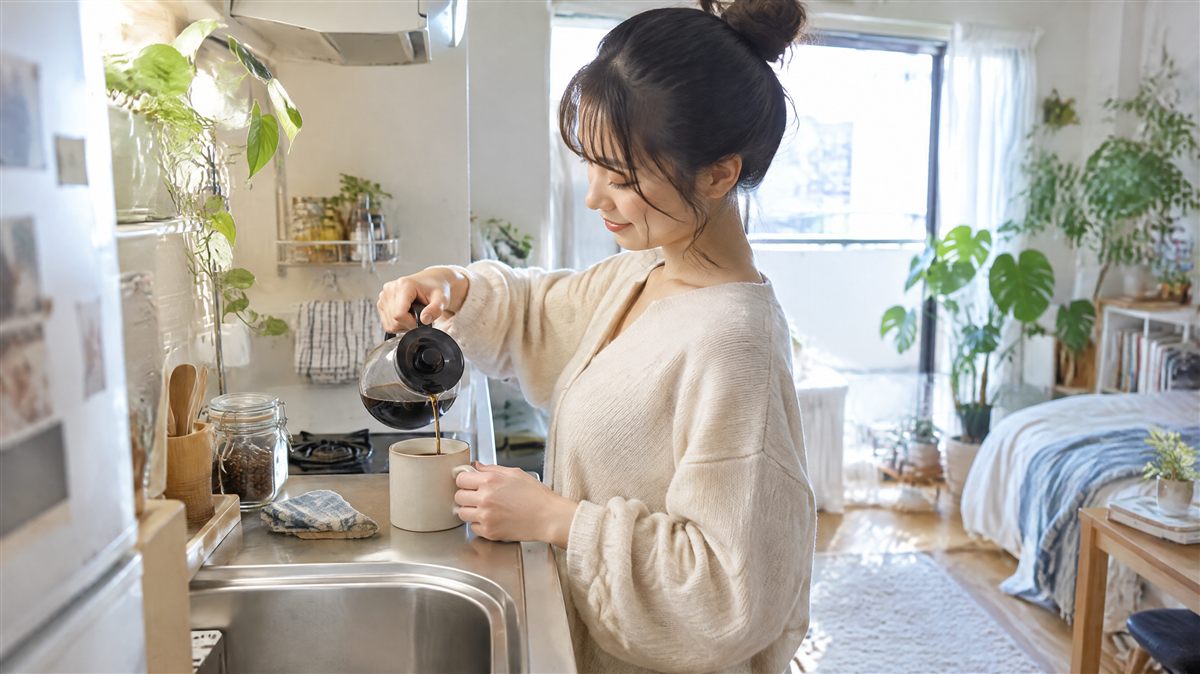 ワンルームのキッチンカウンターでマグカップにコーヒーを注いでいる女性。一人暮らしでお湯をよく使う暮らしのイメージ