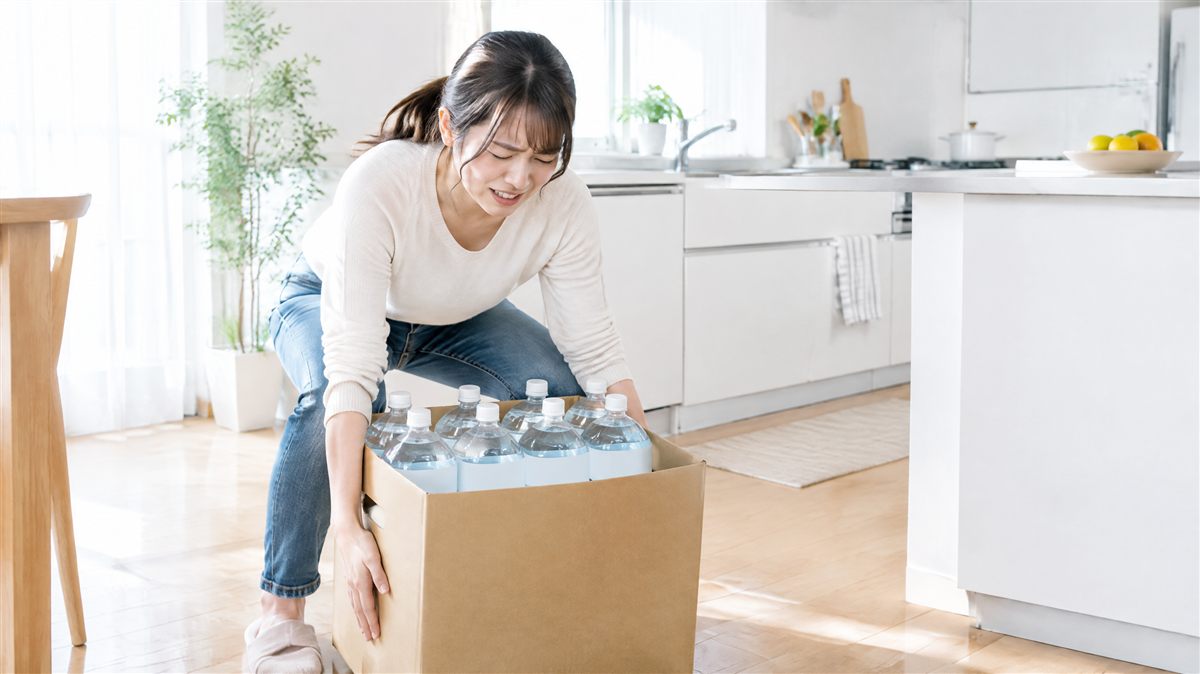 キッチンで2リットルのペットボトル入り飲料水の箱を持ち上げようとしている女性。水の買い出しと運搬の負担のイメージ